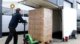Gesloten zakbare enkelasser Humbaur aanhangwagen met geopende achterklep waar een medewerker een pallet met dozen op een palletwagen de laadbak in rijdt