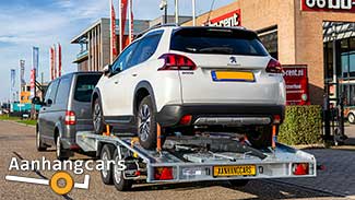 Martz tandemas autotransporter met schuin aflopende achterzijde beladen met een witte personenauto achter een bestelbus